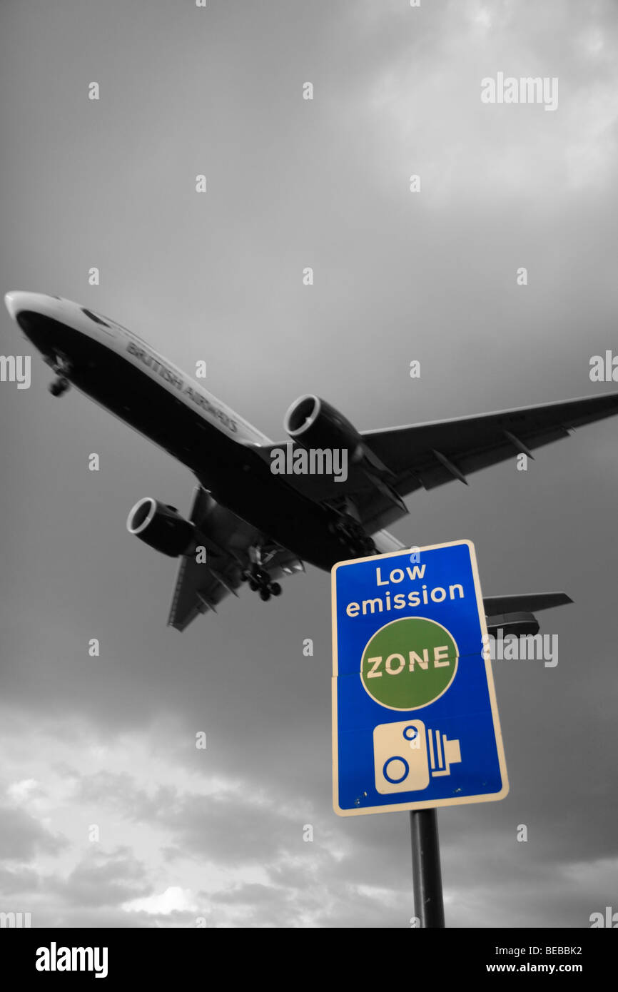 A British Airways areplane passes over 'Low Emission Zone' road sign beside London's Heathrow Airport, UK. B&W & Colour mix. Stock Photo