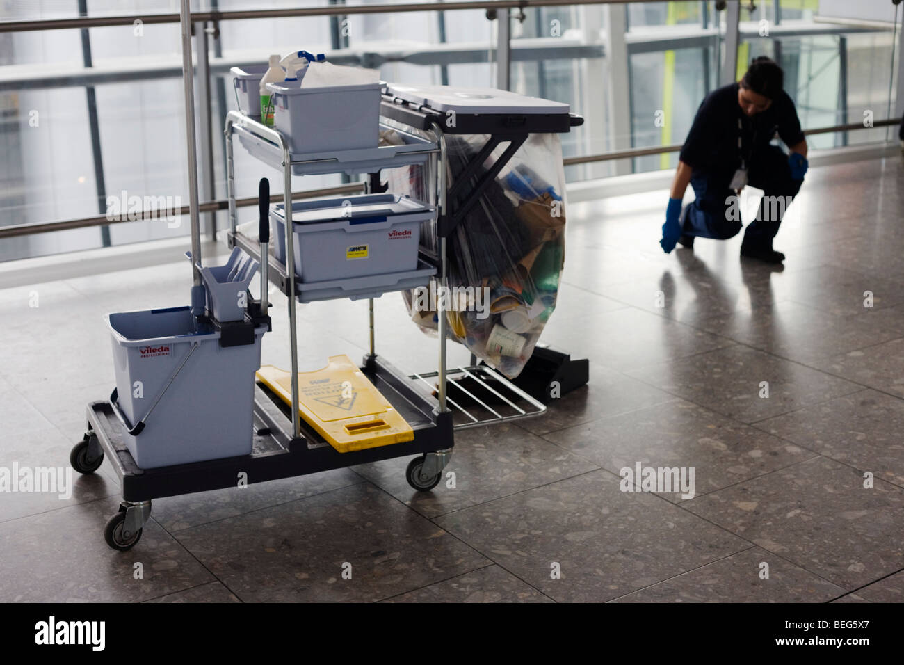 With trolley nearby, a cleaner stoops to remove sticky debris on the concourse floor at Heathrow Airport's Terminal 5. Stock Photo
