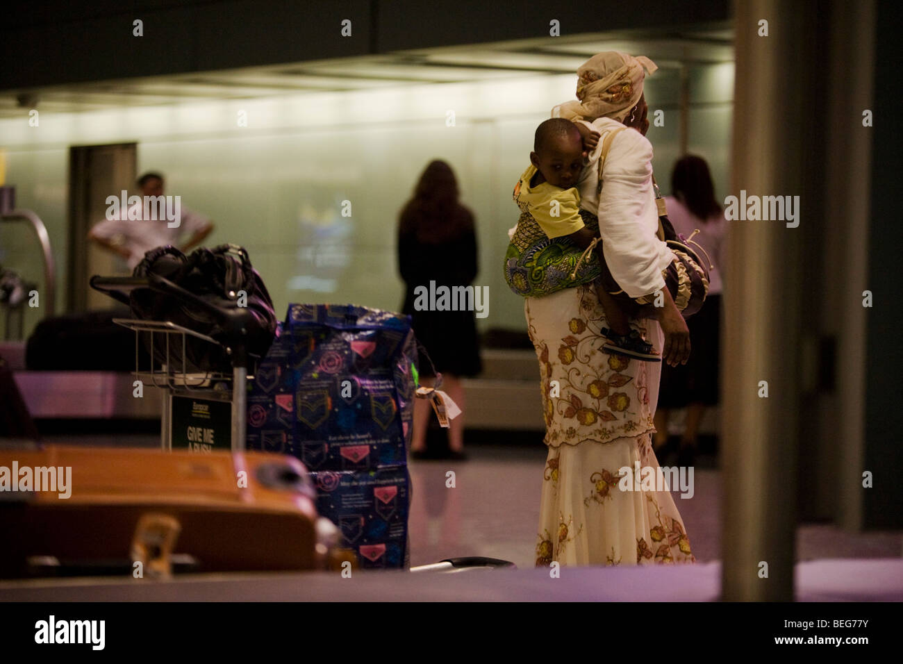 Young child on mother's back await baggage from African flight at Heathrow Airport's Terminal 5. Stock Photo