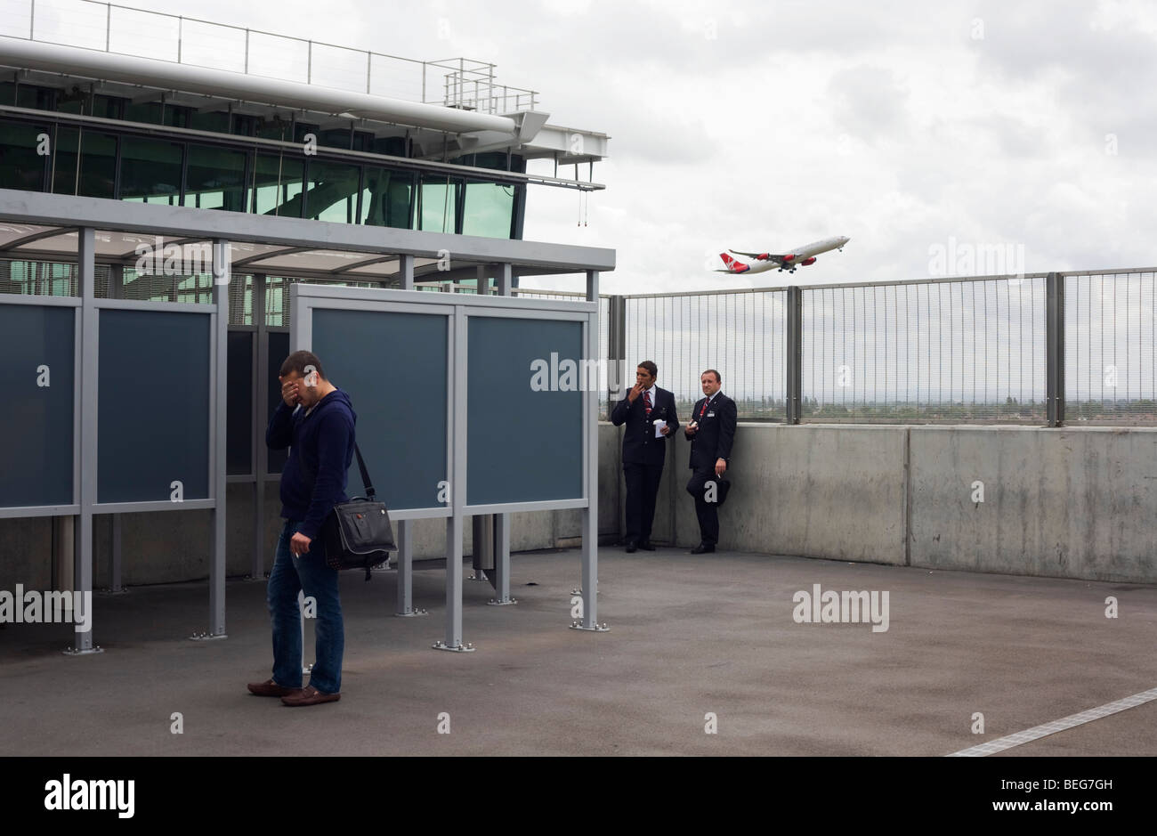 As a Virgin jet takes off overhead, airline employees stop for a cigarette break near the smoking shelter at Heathrow's T5. Stock Photo