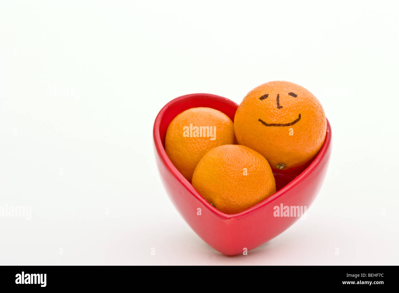 Red heart shaped bowl filled with ripe baby oranges, one with a smiley face. Stock Photo