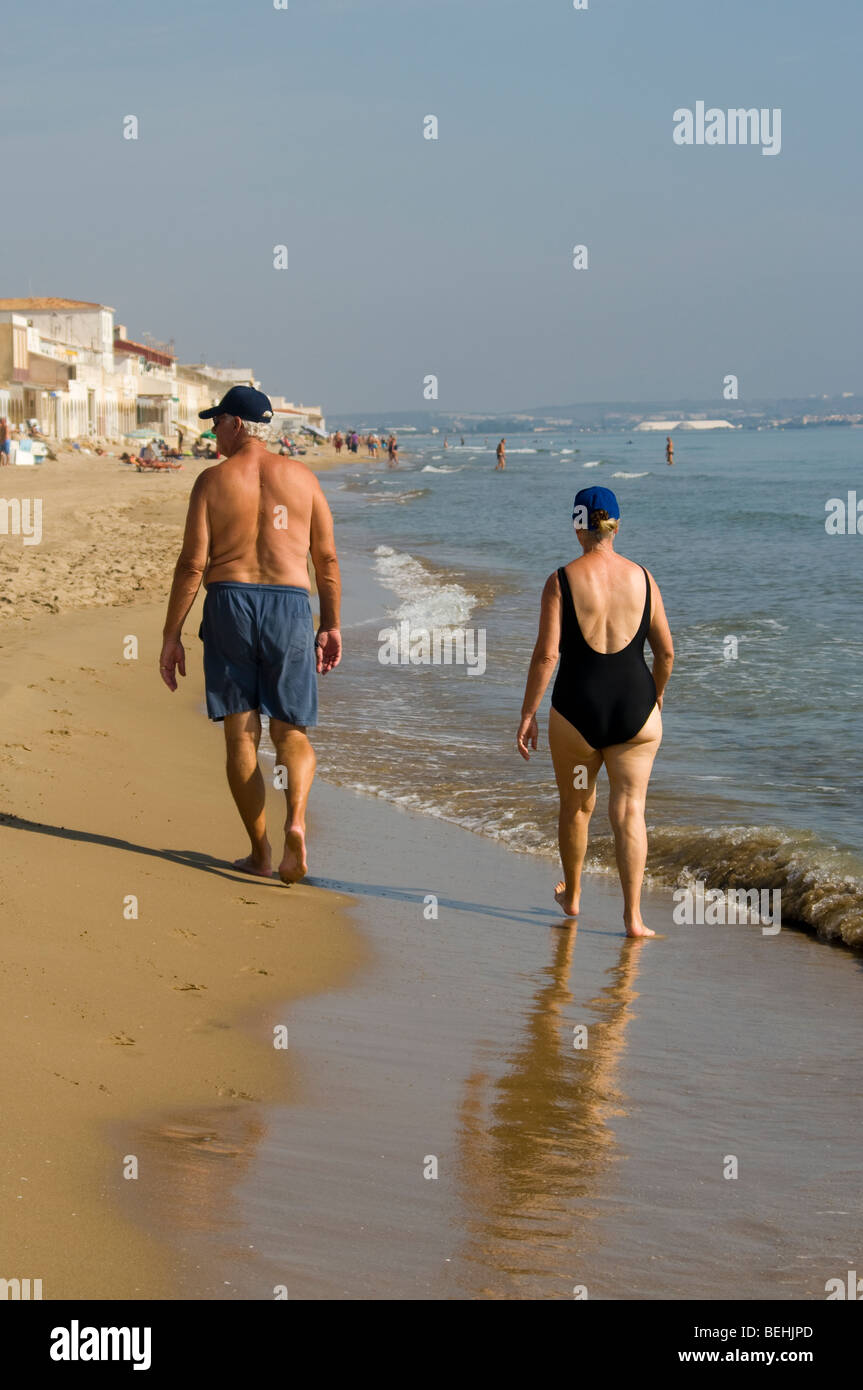 Elderly Couple In Beachwear Walking Down The Beach Seen From Behind Stock Photo