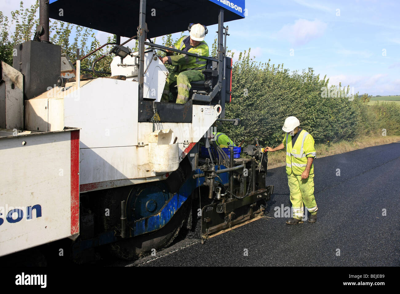 Road surfacing Workmen wearing a yellow hi-vis jacket trousers and white helmet using a tarmac laying machine Stock Photo