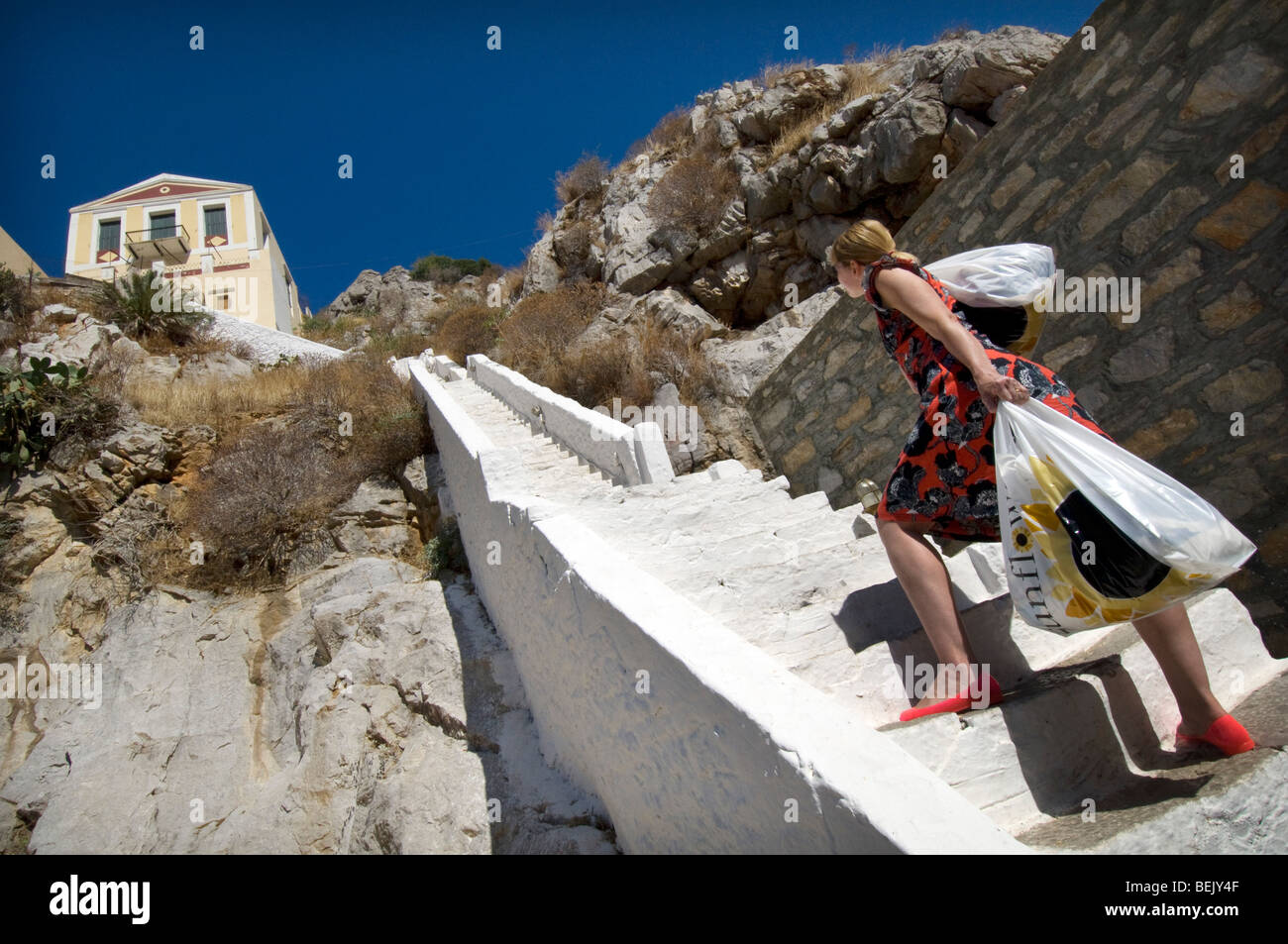 A woman with two bags of heavy shopping climbs steep steps to a house on the Greek island of Symi Stock Photo