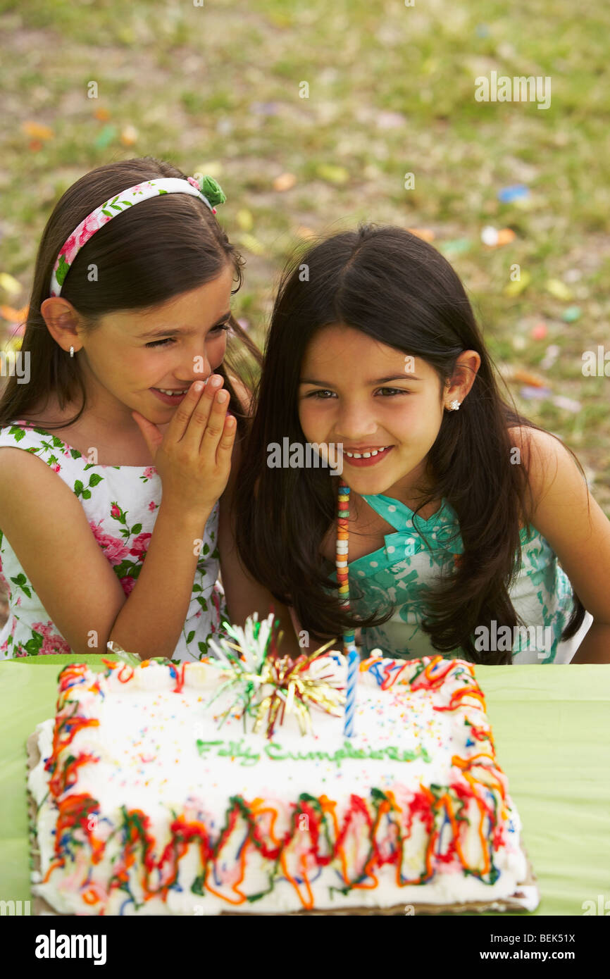 Girl sitting in front of a birthday cake and whispering to her friend ...