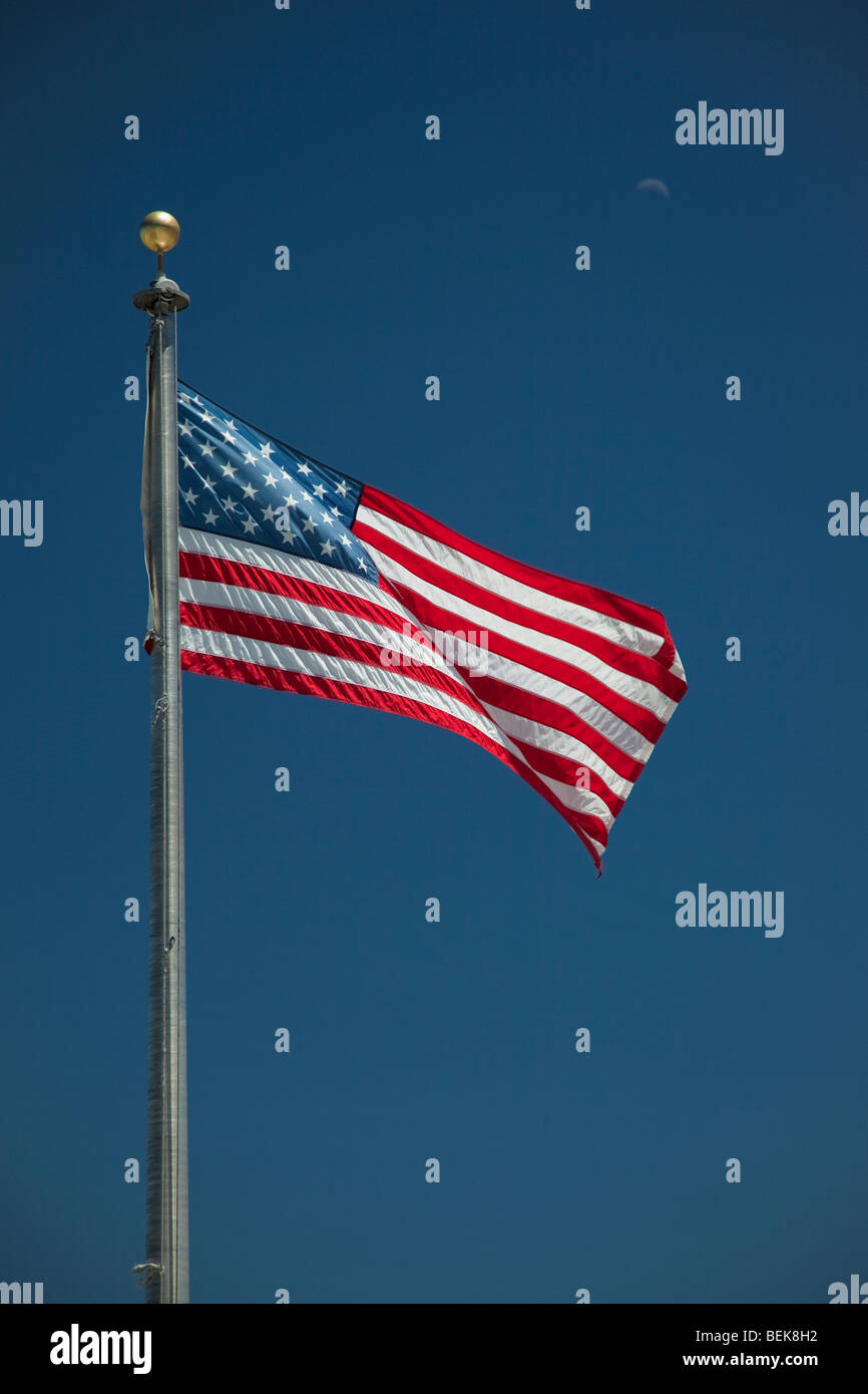 Sunlit American flag against a clear blue sky with a crescent moon Stock Photo