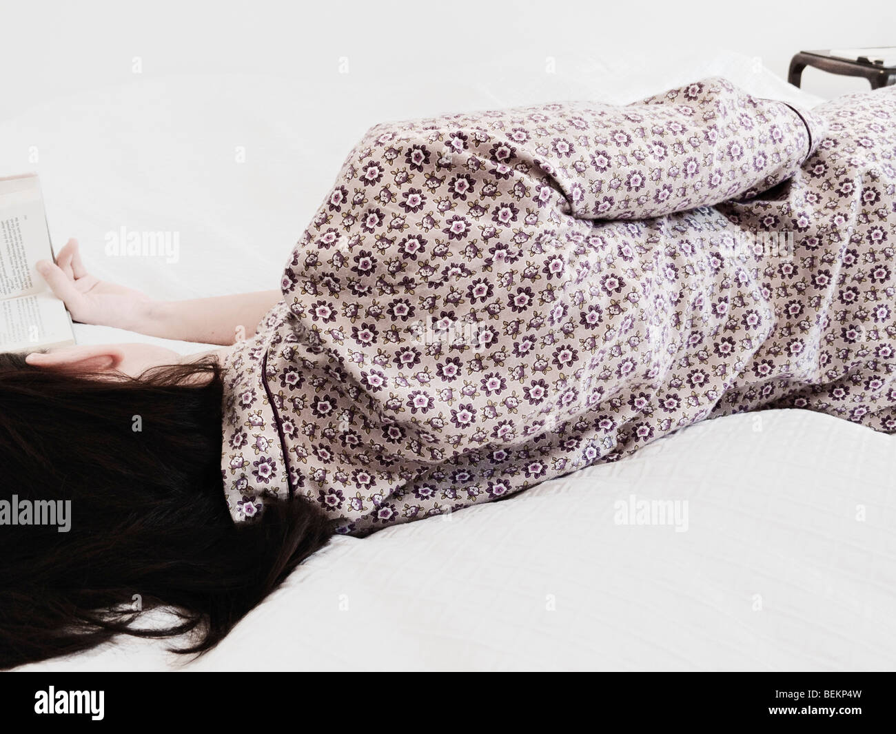 Young Woman Wearing Pajamas Relaxing On Bed, Reading A Book Stock Photo