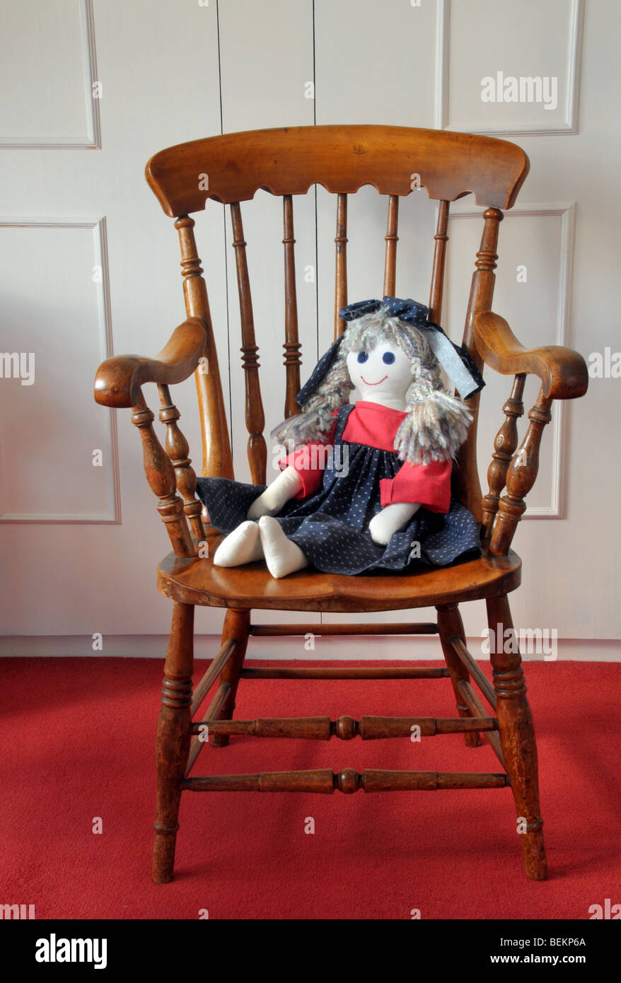 A home made cloth doll dressed in a blue and red pinafore  with a bow in her hair, sits on a wooden chair. Stock Photo