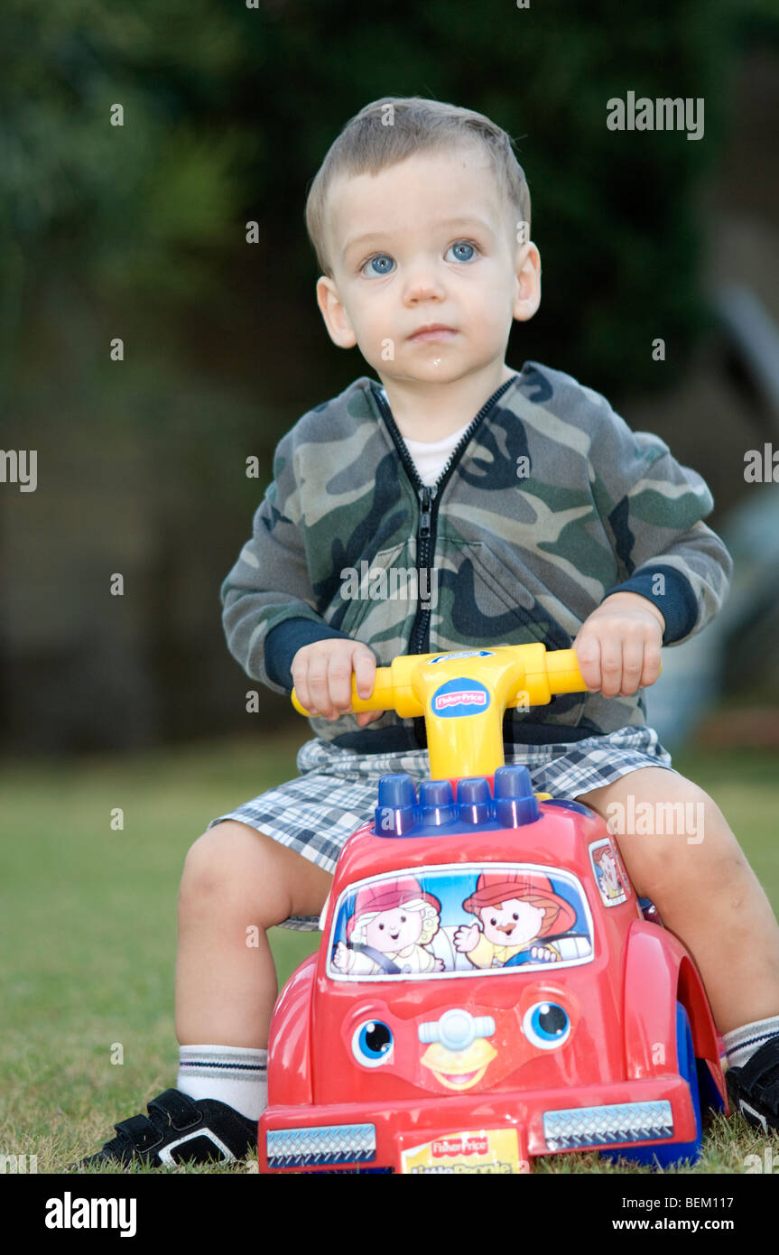 A blue-eyed young boy about 1 year old rides atop a toy firetruck wearing a camouflage pattern jacked and short pants.  Outdoor. Stock Photo