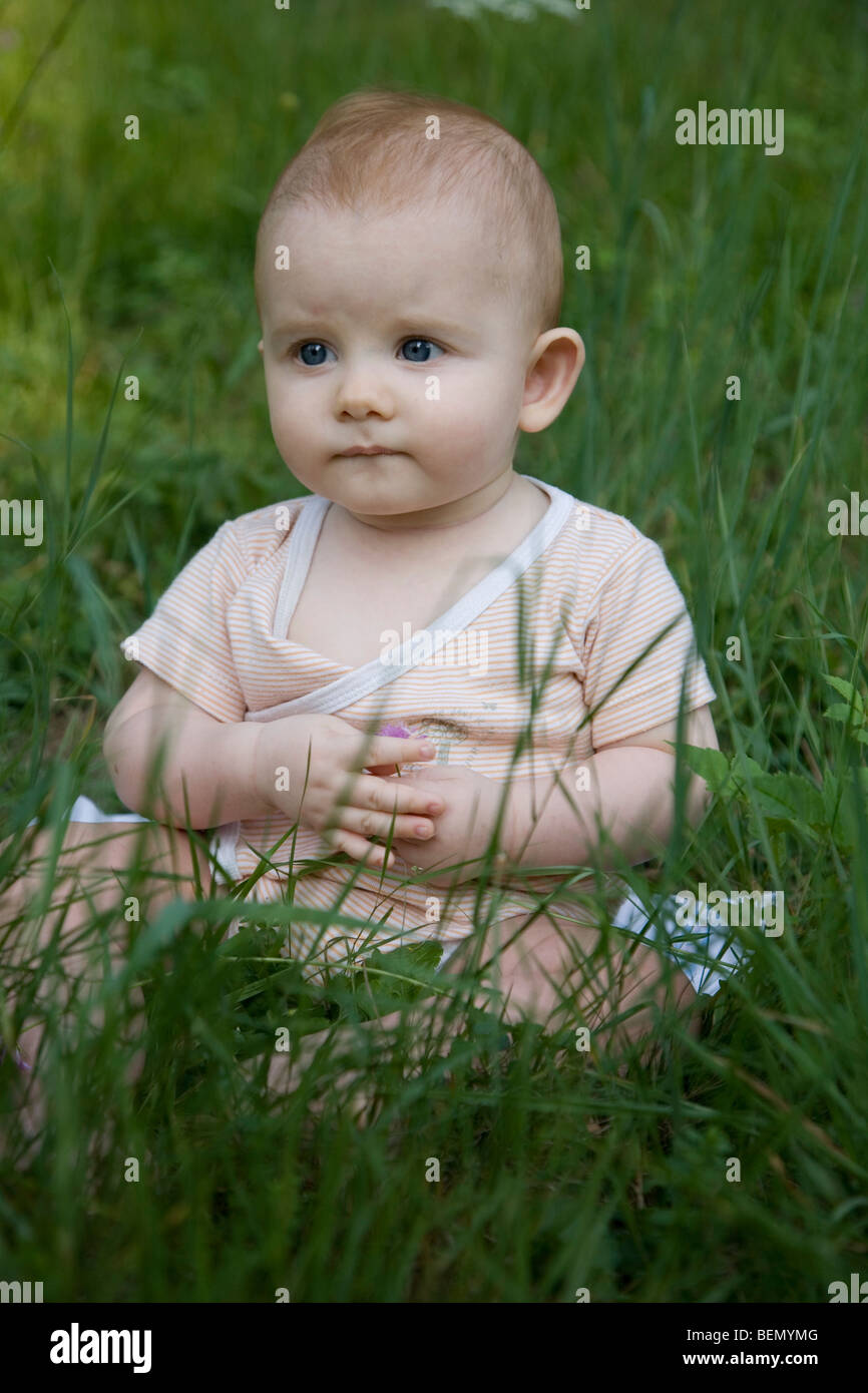 eight-months old strawberry-blond baby sitting on the meadow, observing doubtfully and a little timorous Stock Photo