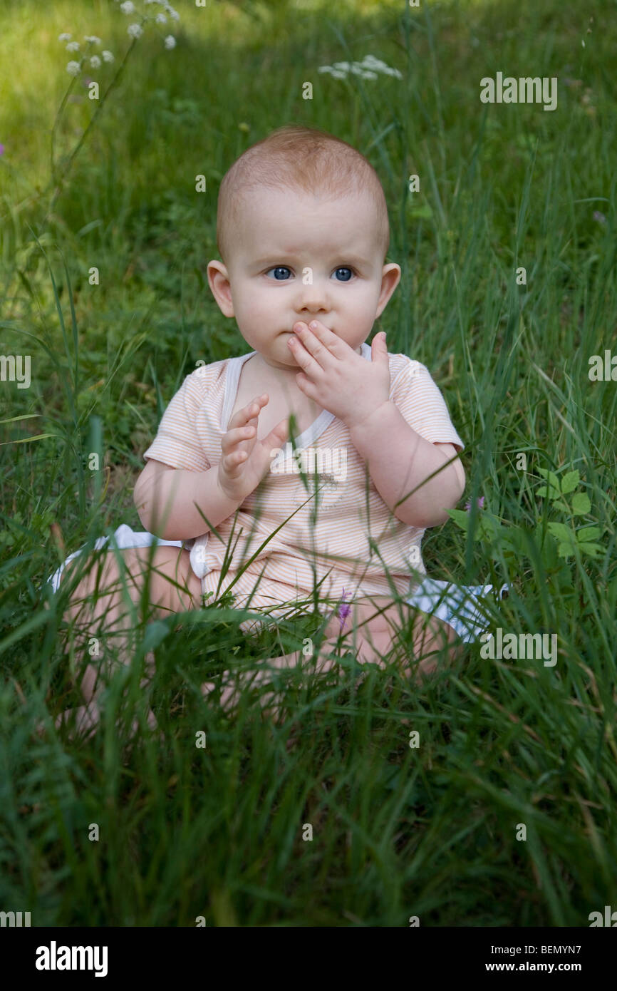 eight-months old strawberry-blond baby sitting on the meadow, looking doubtfully Stock Photo
