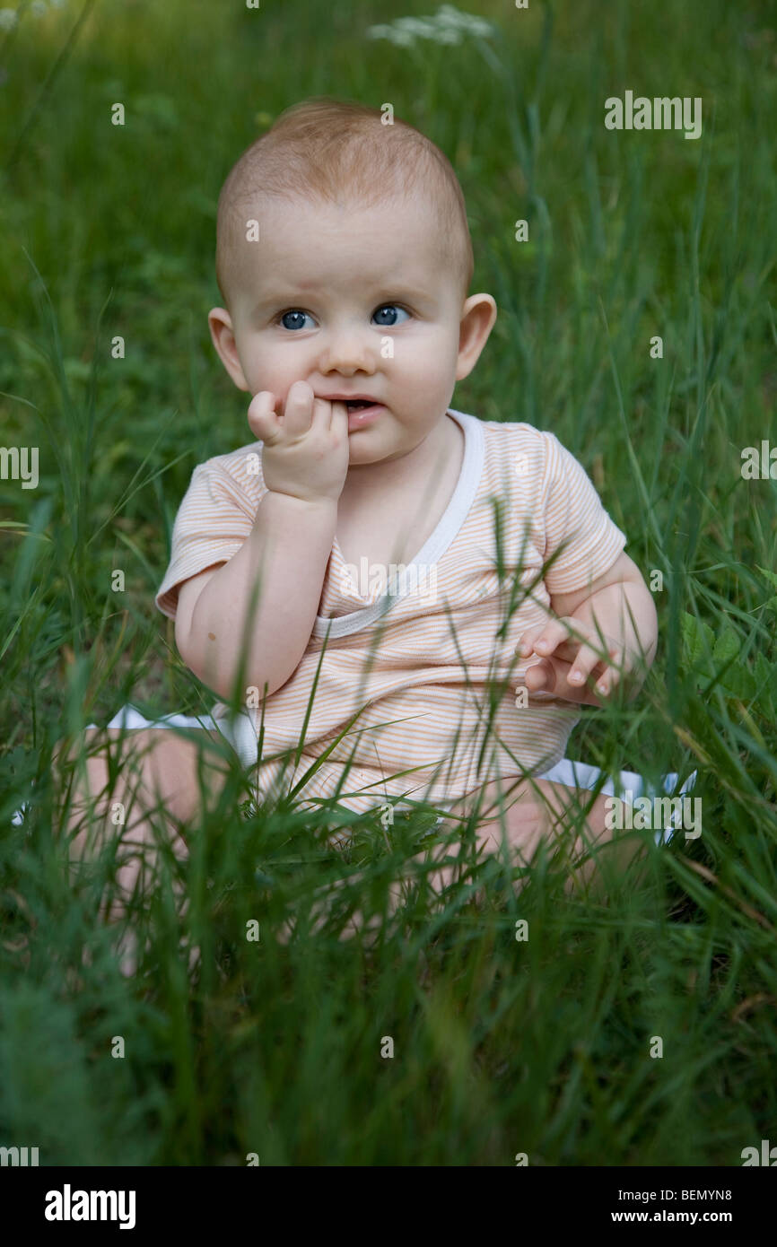 eight-months old strawberry-blond baby sitting on the meadow, observing dubiously and biting on his fingers Stock Photo