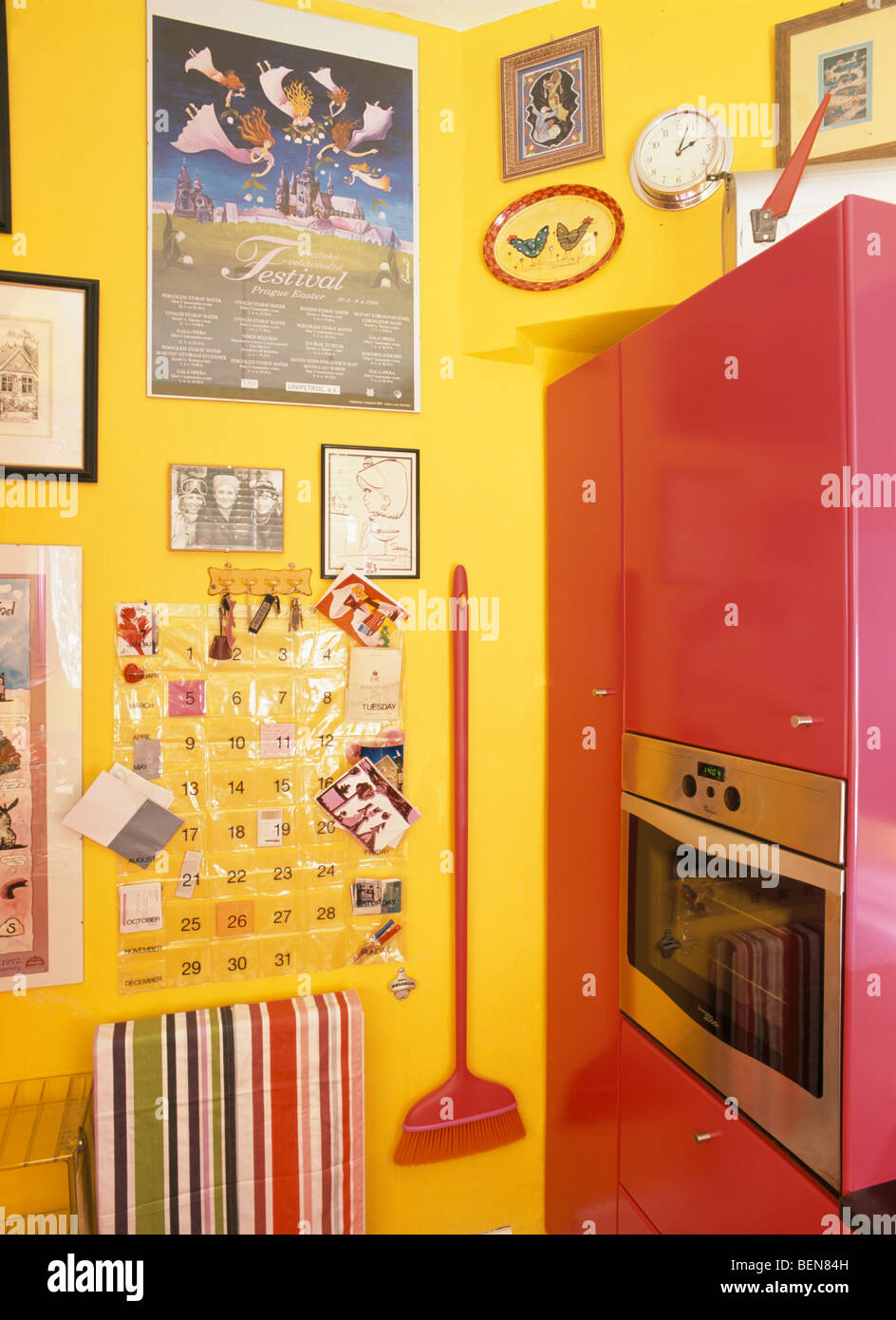 Pictures and red broom on wall beside oven in fitted pink unit in bright yellow kitchen Stock Photo