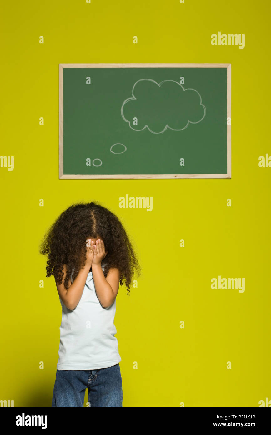 Little girl covering face with hands, thought bubble on chalkboard behind her Stock Photo
