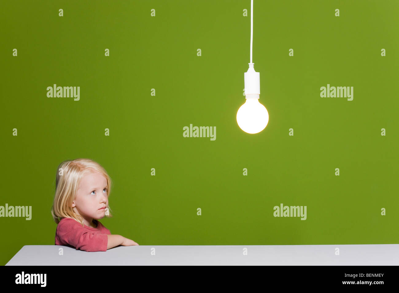 Little girl biting lip looking with concern up at illuminated light bulb suspended overhead Stock Photo