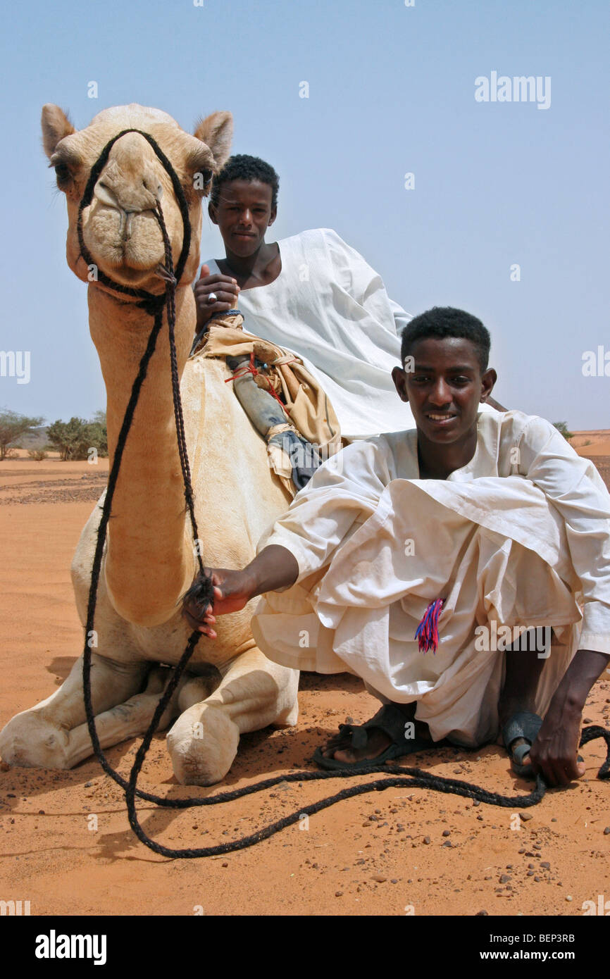 Two Nubian boys dressed in thawbs with dromedary camel (Camelus dromedarius) in the Nubian desert of Sudan, North Africa Stock Photo