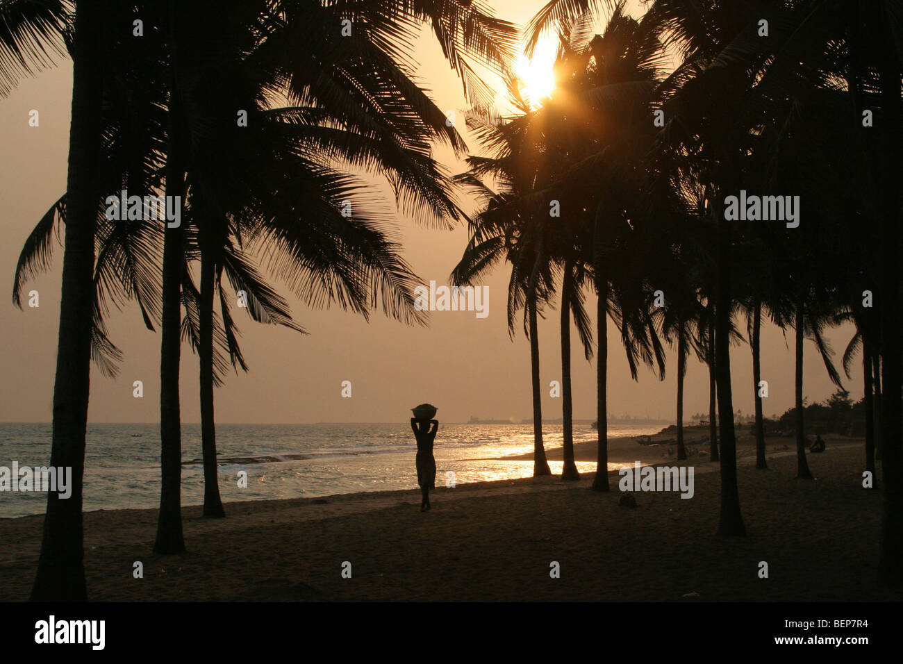 Silhouetted palm trees and woman carrying merchandise on her head on beach at sunset, Gulf of Guinea, Lomé, Togo, West Africa Stock Photo