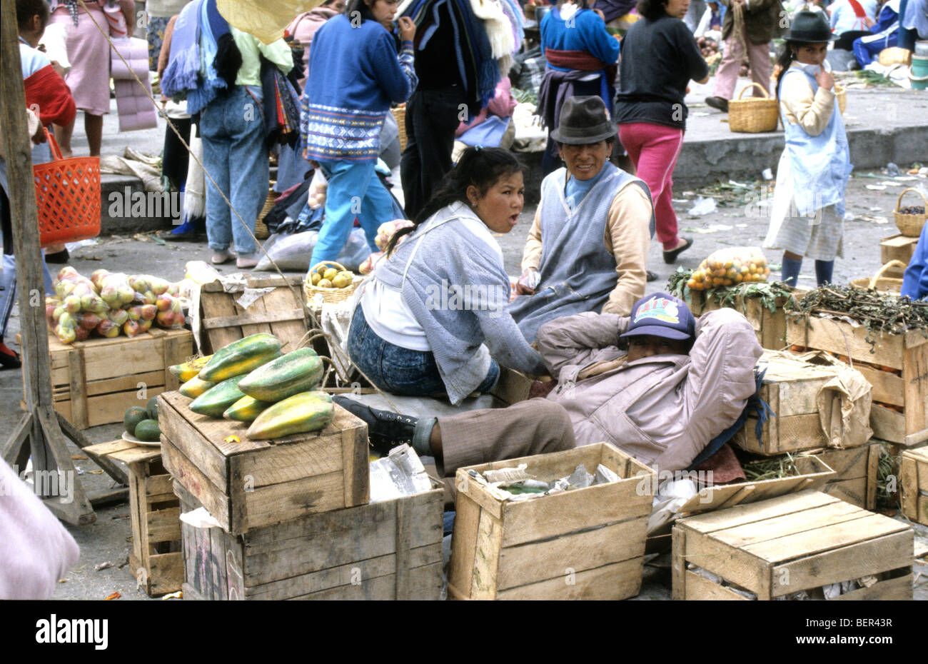 Two women sit up amongst small wooden boxes surrounded by fruits for sale.  Local upland Ecuador market. Stock Photo