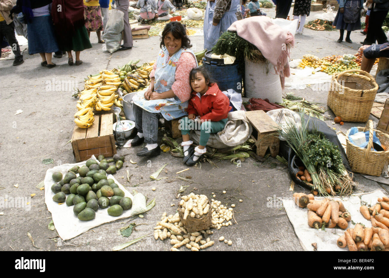 Smiling woman and child sitting on small wooden box amidst fruits and vegetables. Local upland Ecuador market. Stock Photo