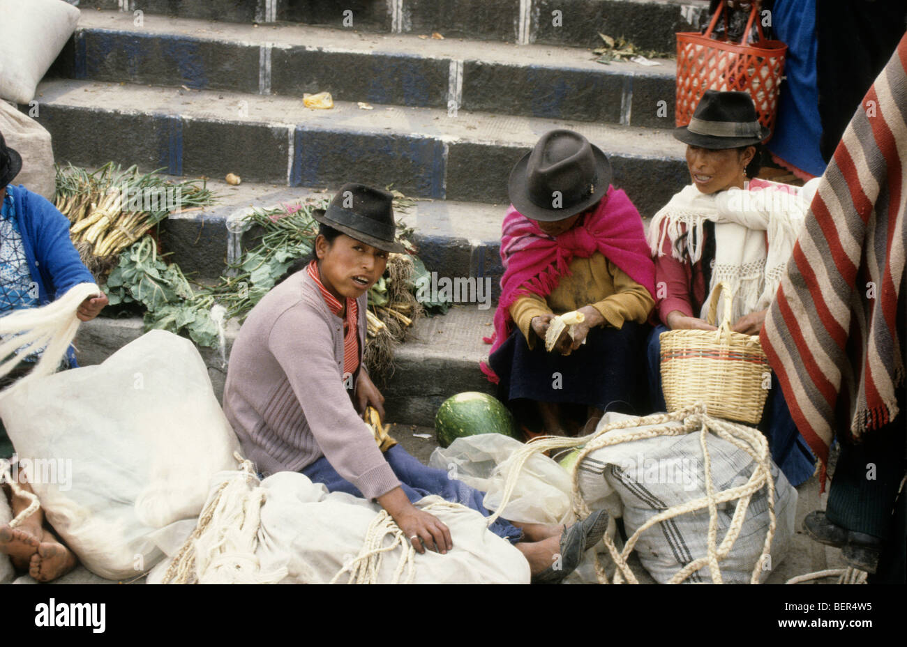 Three women sitting at base of steps amidst sacks of vegetables. Ecuador highlands local market. Stock Photo