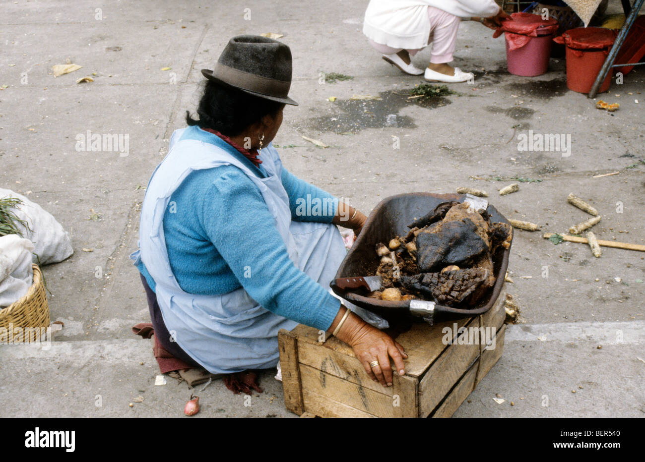 Woman squatting before a upturned wooden box with a metal tray of slabs of dried cooked meat.  Ecuador highlands local market. Stock Photo
