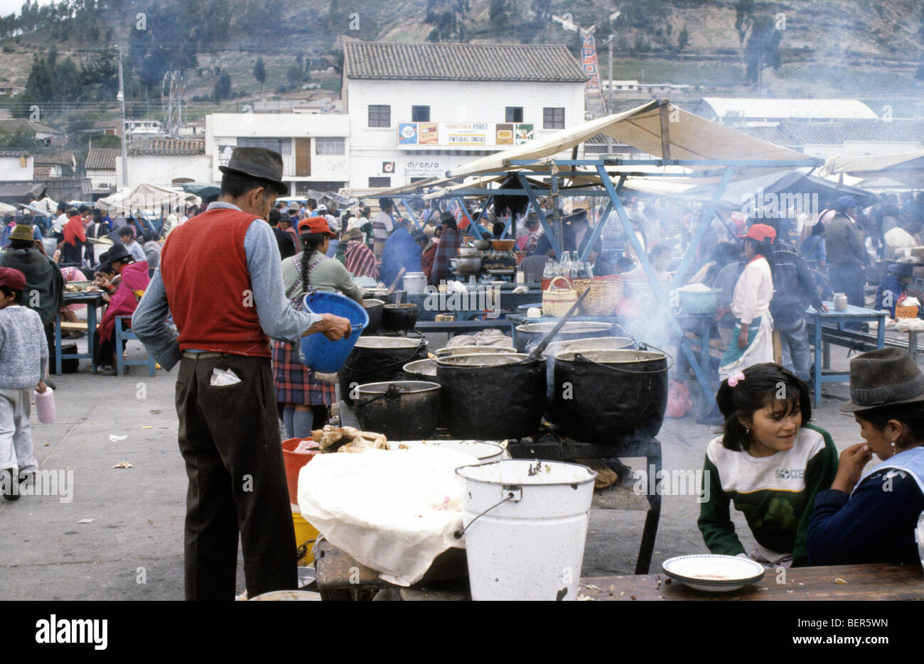 Male stall owner hold up plastic bucket and surveys line of deep metal cauldron like pots on cooked food stall.  Ecuador market Stock Photo