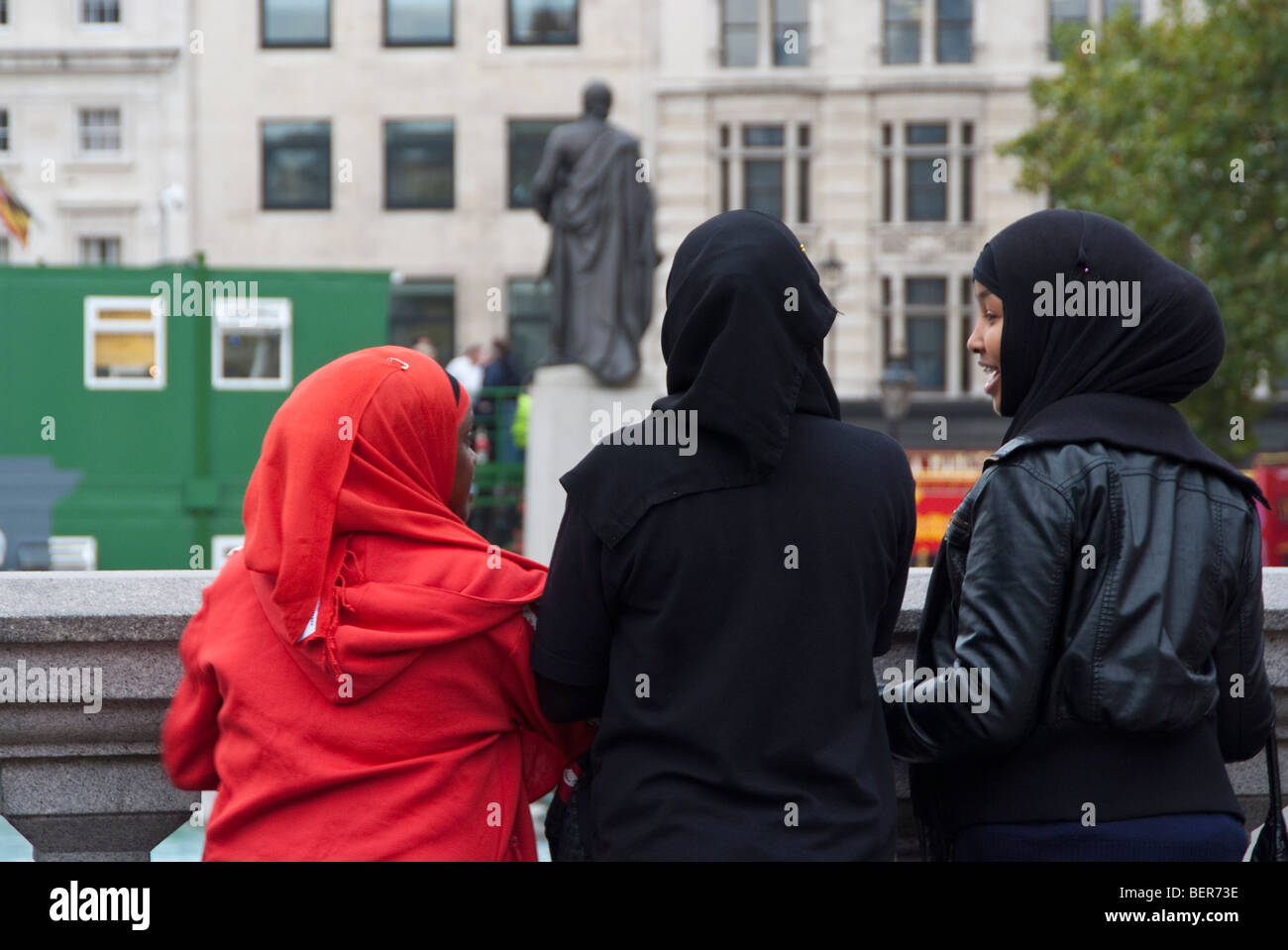 Three muslim girls, two wearing back headscarfs and one a bright read headscarf, look out over Trafalgar square in London Stock Photo