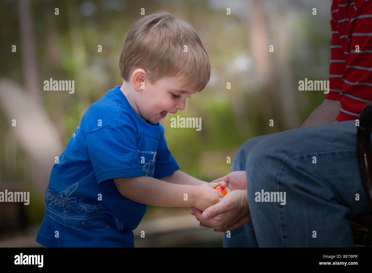 Happy excited 2-3 year old blond Caucasian boy outdoors, hands in dads cupped hands opening Easter egg shallow depth of field Stock Photo