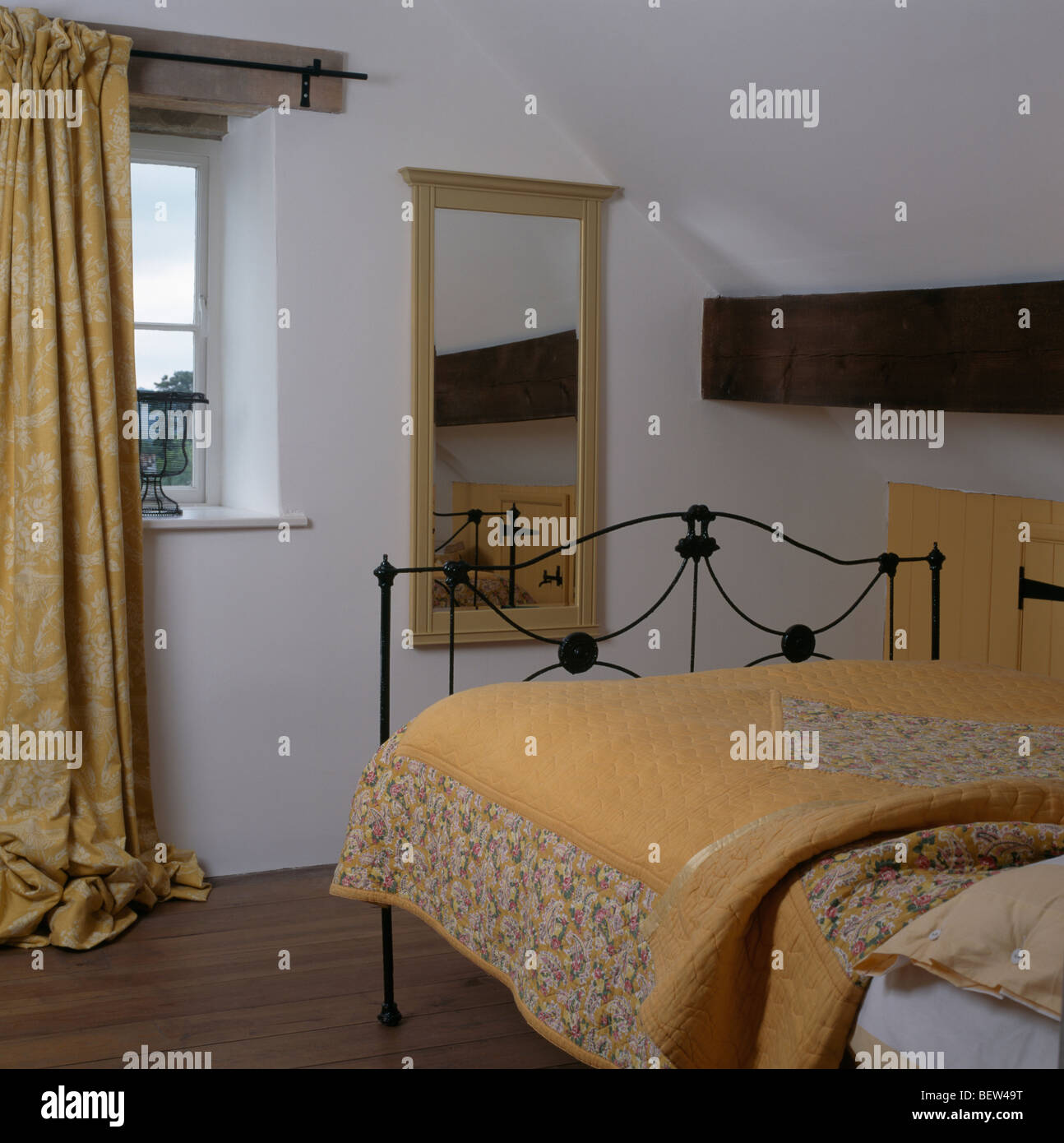 Yellow quilt on iron bed in white cottage bedroom with patterned yellow curtain at window Stock Photo