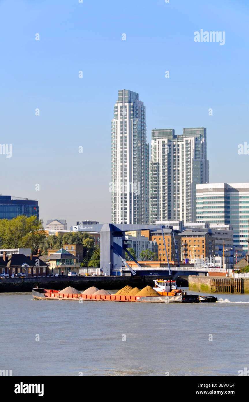 Tug boat and barge loaded with construction aggregates at entrance to London Docklands waterways apartment blocks beyond Stock Photo
