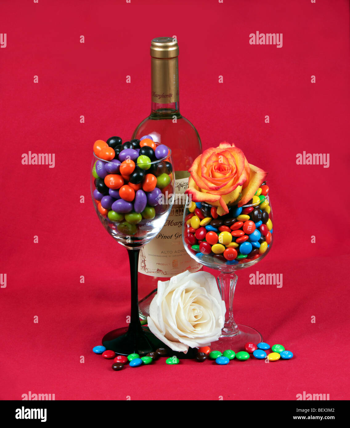 A bottle of wine two wineglasses filled with M&M candy and two beautiful roses. Shot against a red background. A holiday shot. Stock Photo