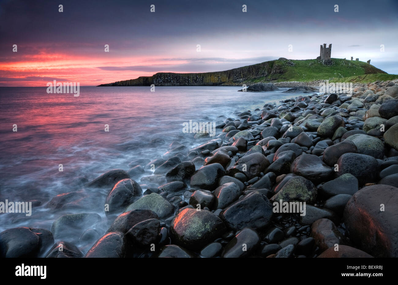 The pink pre-dawn light illuminates the sea and Graymare rocks at Dunstanburgh Castle. Stock Photo