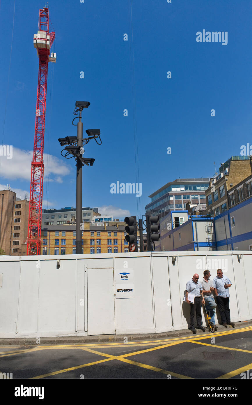 Cluster of seven CCTV / Congestion Charge cameras overlook three men standing in front of a white hoarding (of a building site). Stock Photo