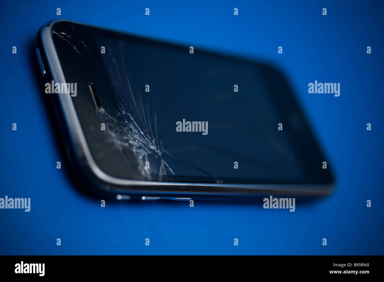 A damaged Apple iPhone device placed on a blue backround.  The main control screen has been cracked at the top. Stock Photo