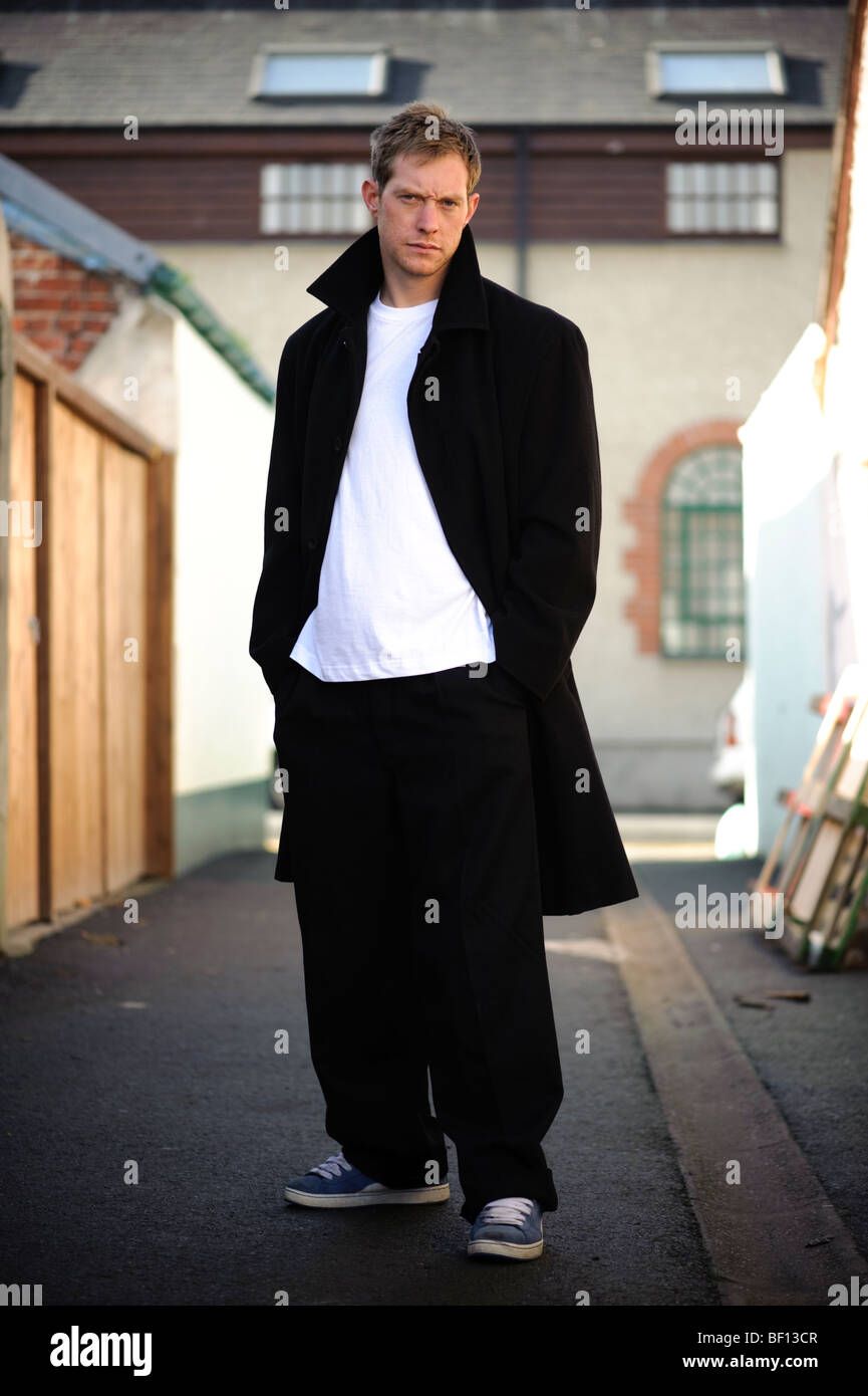 30 year old man standing alone in an alleyway with his hands in his pockets wearing black coat white t-shirt sneakers, uk Stock Photo