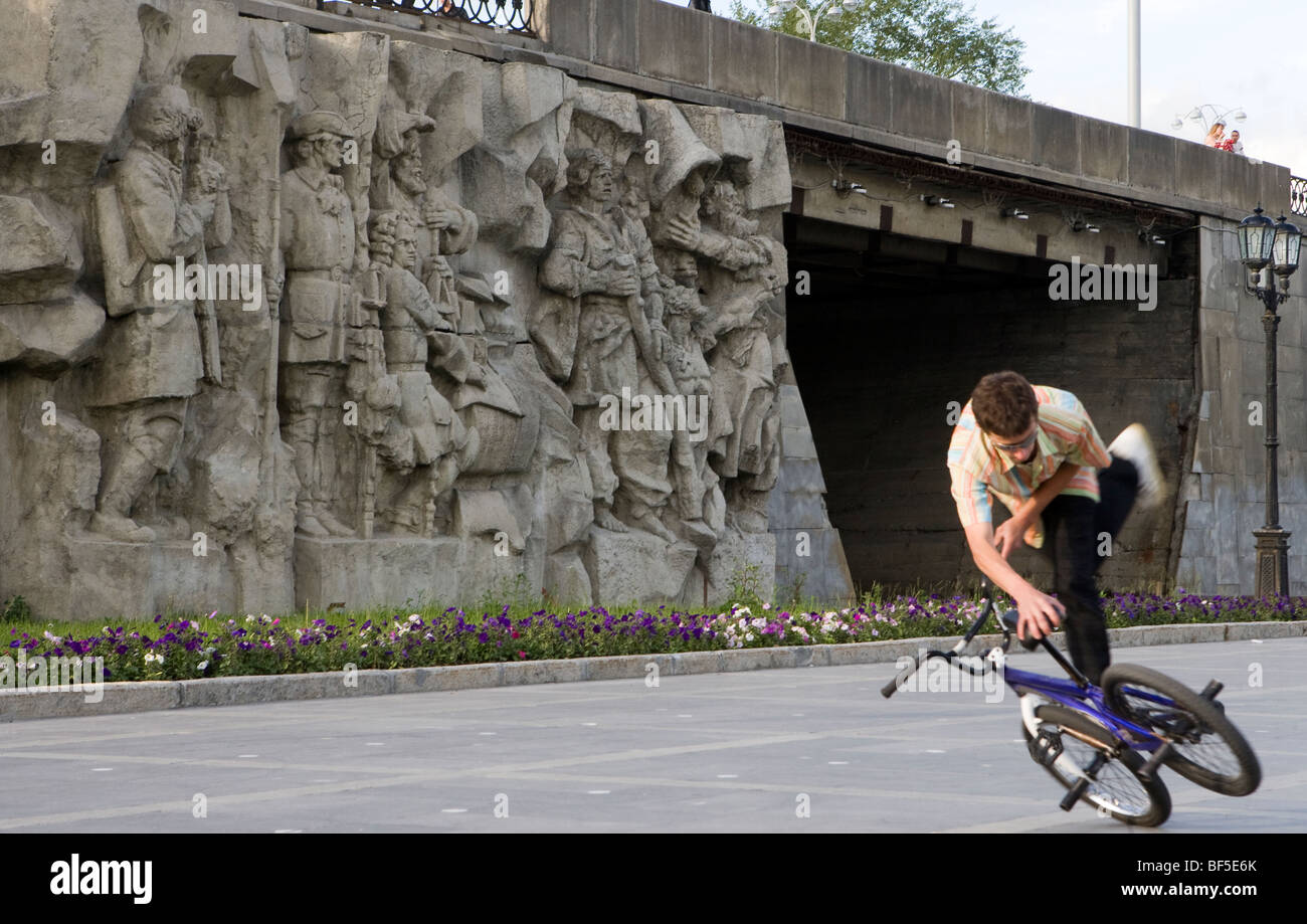 Teenage boy doing bicycle stunt near bridge. Monument bas-relief birth of the city, Yekaterinburg, Russia Stock Photo