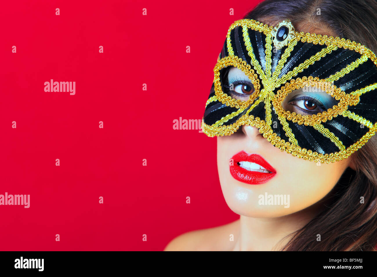 Beautiful brunette woman wearing a black and gold masquerade mask and bright red lipstick against a red background. Stock Photo