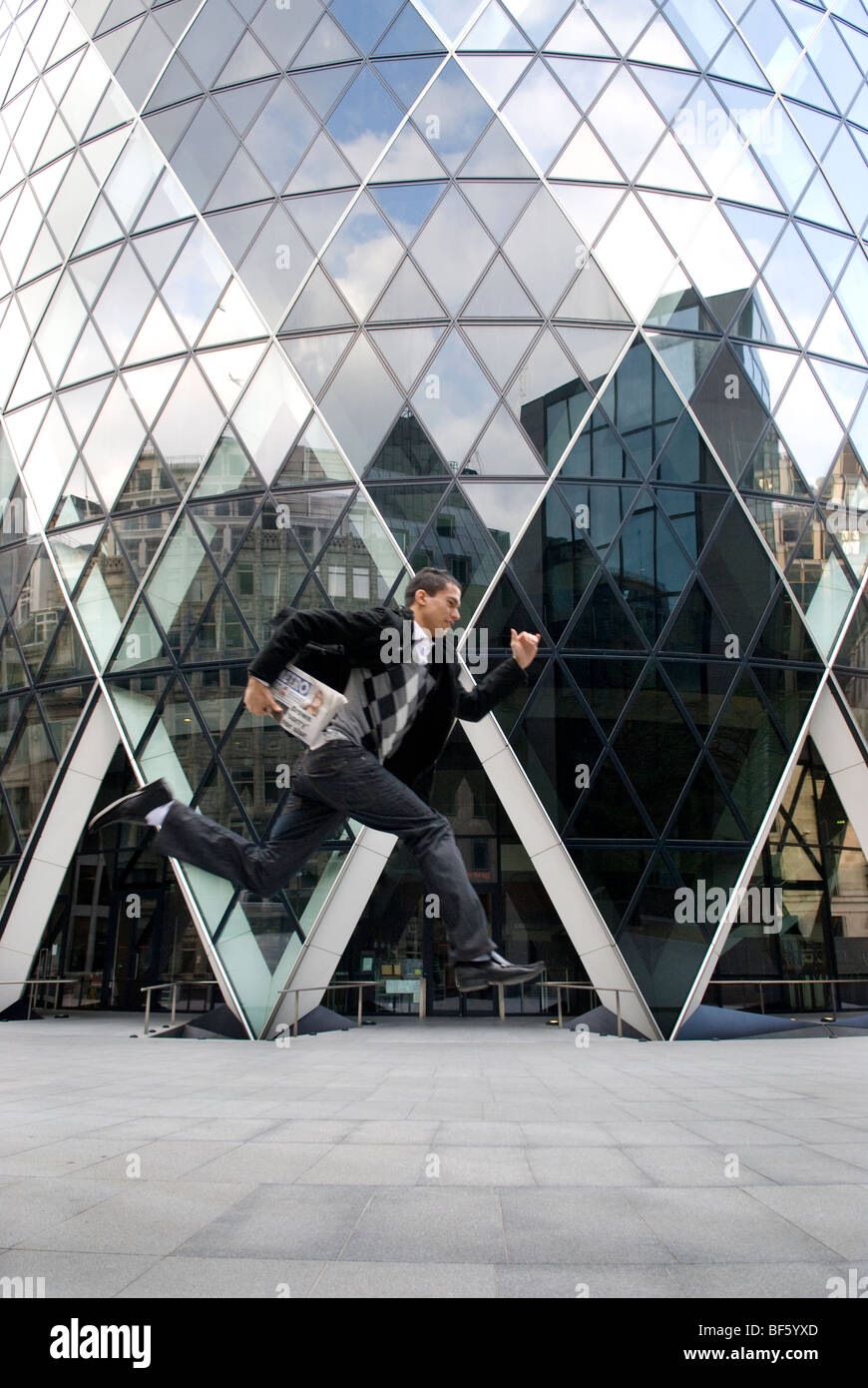 A suited young man is leaping in front of The Gherkin, St Mary's Axe, London, holding a newspaper in his hand Stock Photo
