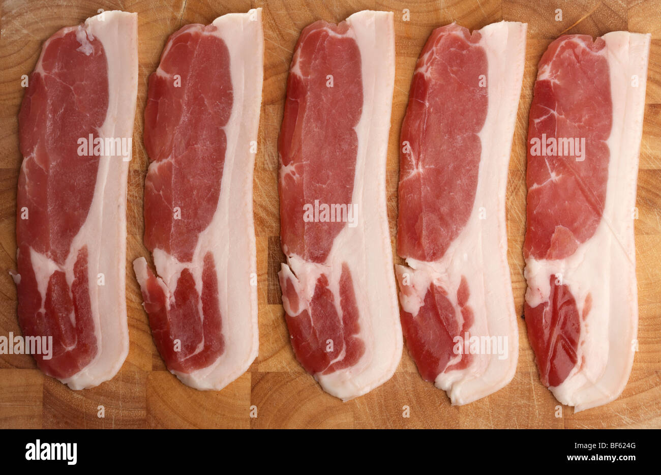 five strips of raw back bacon from organic british saddleback pigs reared in ireland laid out on a wooden butchers block Stock Photo