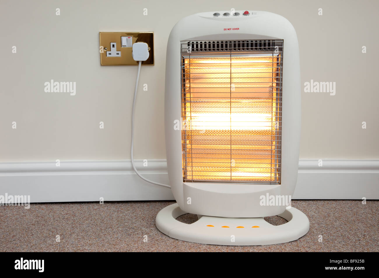 Electric halogen room heater with three heat settings plugged into electrical wall socket and switched on with low setting. UK Stock Photo