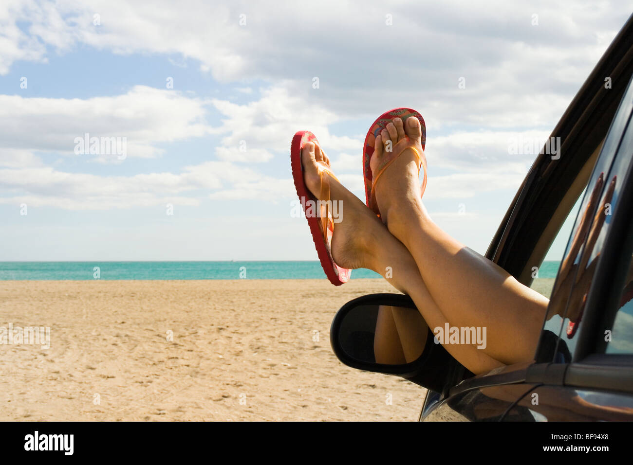 Woman's legs and feet in orange flip flops sticking out of car window in front of empty beach Stock Photo
