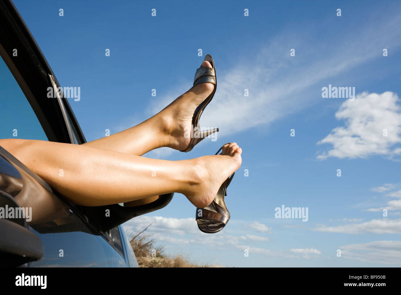 Woman's legs and feet wearing silver high heeled shoes sticking out of car window. Stock Photo