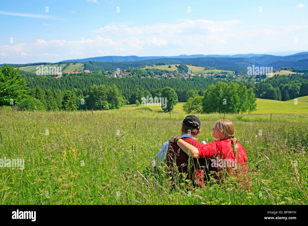 A couple sitting in high grass enjoying the panoramic view of St. Andreasberg, Harz Mountains, Lower Saxony, Germany Stock Photo