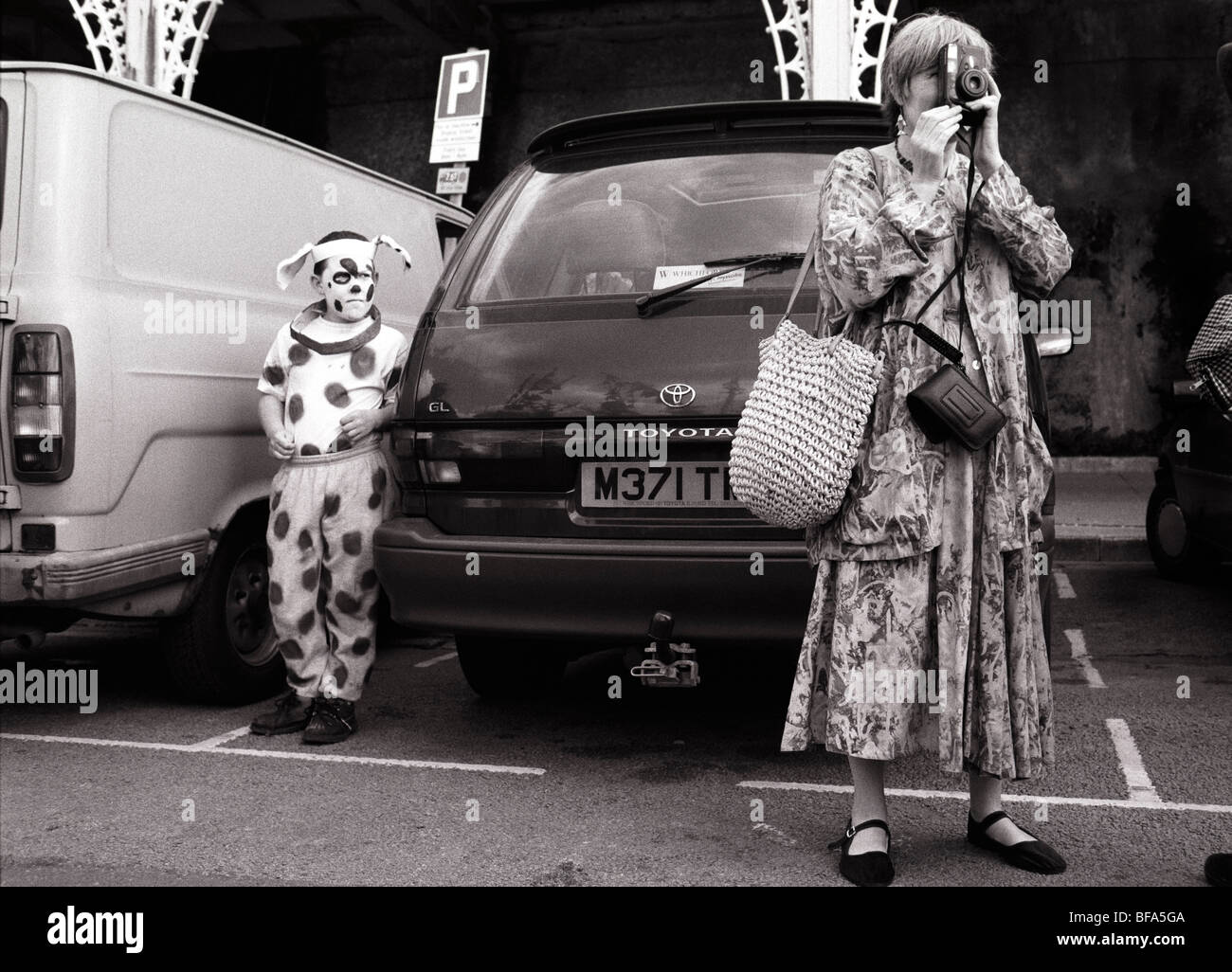 A boy in a dalmation dog fancy dress parade in Brighton waits by a car as his mother takes a photograph. Stock Photo