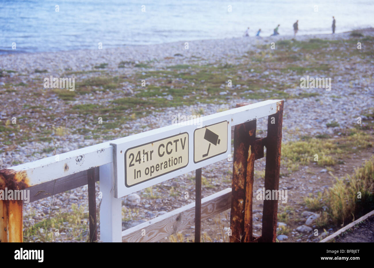 Rough stony beach with weeds and sea beyond with rusty gate and iconic image of camera and warning 24hr CCTV in operation Stock Photo