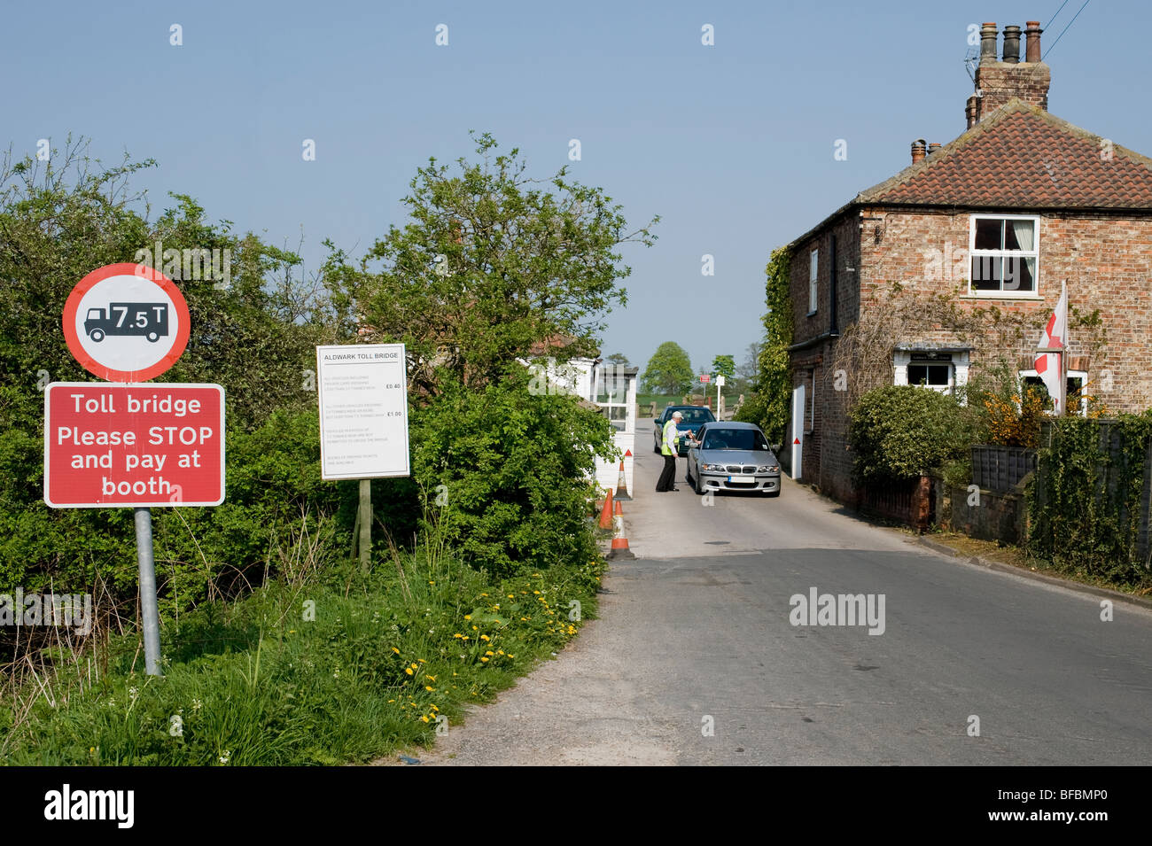 Car driver paying at Aldwark toll bridge, handing money to man on duty by booth. (Clear signage & toll keeper's house) - North Yorkshire, England, UK. Stock Photo