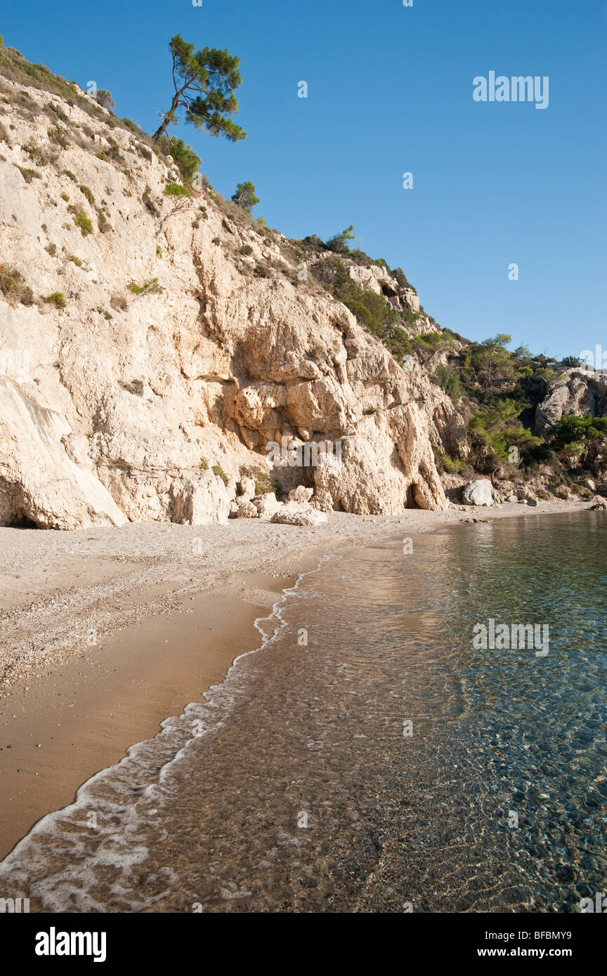 View along a small sandy beach on a greek island with a tree and some rocks and cliffs in the background. Stock Photo