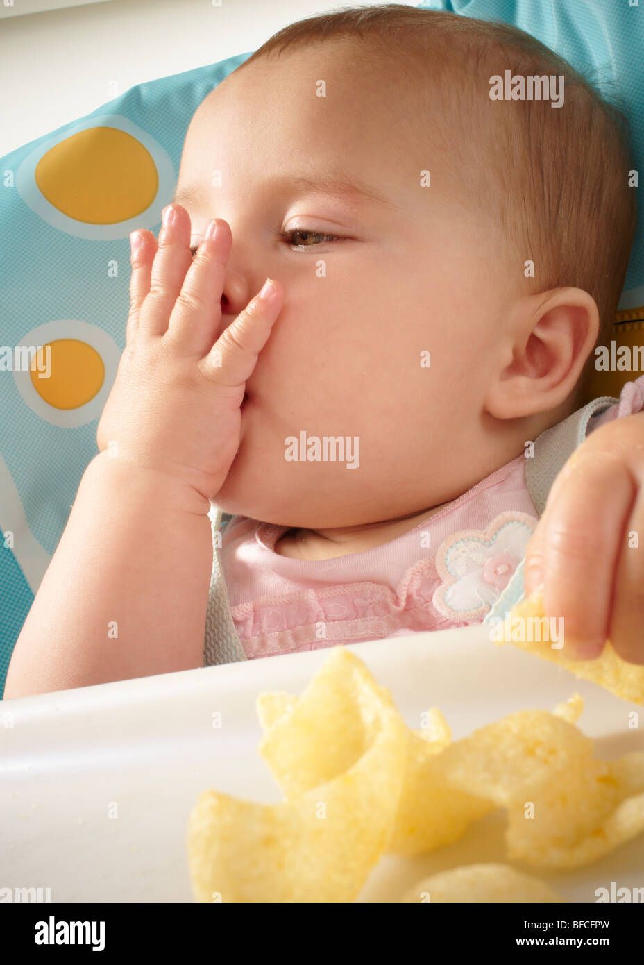 Baby Girl in pink top feeding and being fed in blue and yellow high chair Stock Photo