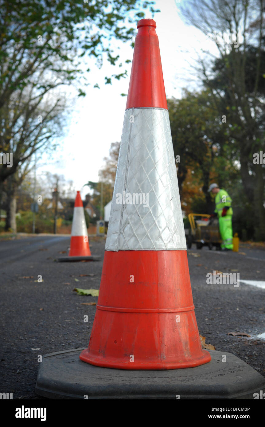 A red and white cone used to direct and contain road traffic in the UK. In the background is a road maintenance worker. Stock Photo