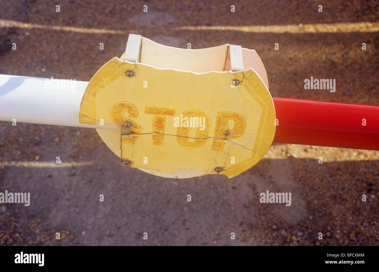 Red and white vehicle barrier across road with strips of sunlight and with a broken split and faded plastic sign stating Stop Stock Photo